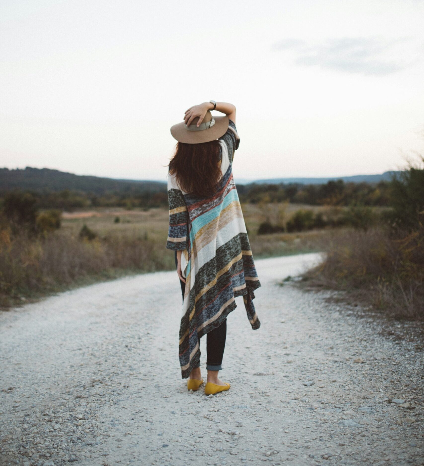 A woman standing looking down a country road wearing a hat and sweater